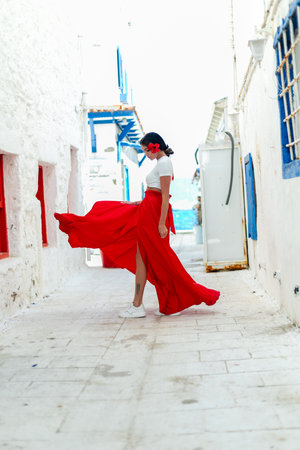Sunny escape: A brunette beauty in a red summer dress strikes a pose against a white wall backdrop with Greek villas, evoking the spirit of travelの写真素材