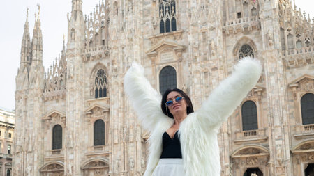 City elegance: A beautiful tourist girl exudes charm in Milan, smiling brightly in her white fur coat against the backdrop of Milan Cathedralの写真素材