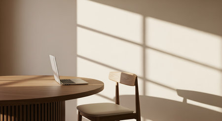 A minimalist workspace with a laptop on a round wooden table, an empty chair beside it. Golden afternoon sunlight bathes the scene, casting soft shadows and creating a peaceful ambの素材