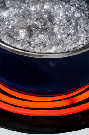 Vertical close-up shot of a blue pot on a stove top on a red hot burner holding boiling water.  Only...