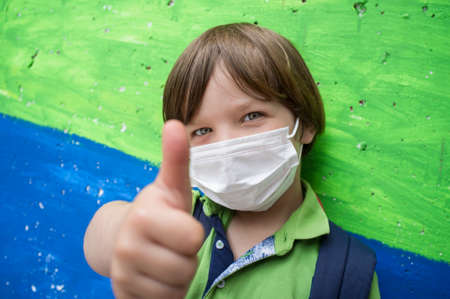4 years little boy wearing a face mask doing the thumbs up gesture. Lively colored concrete block as...
