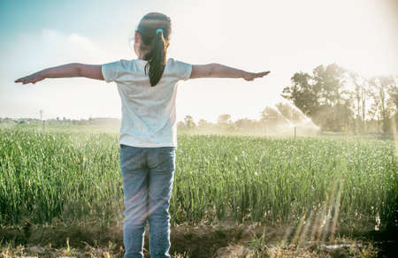 Child girl under sprinklers at work with backlit sunrays at onions field. Guadiana meadows, Badajoz,...