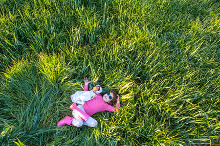 Little girl and her doll lying on green cereal field at sunset, Spain