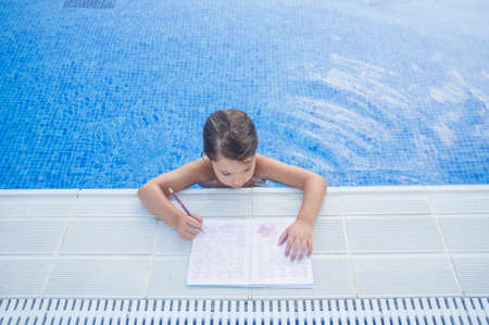 Child girl doing holidays homework over swimming poolside. Summer homework concept for kidsの写真素材