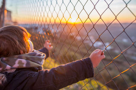 Kid boy holding hands on wire fence at sunset. Concept for child freedom, abuse, human trafficking,...