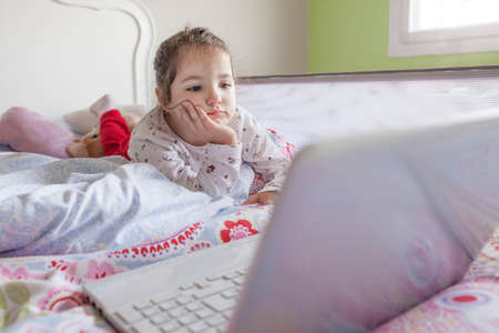 Little girl lying in bed and watching cartoons with a laptop in her bedroom. She looks entertained