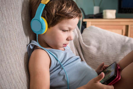 4 year old boy in headphones watching videos with smartphone. Enjoying sitting on sofa in living room at homeの写真素材