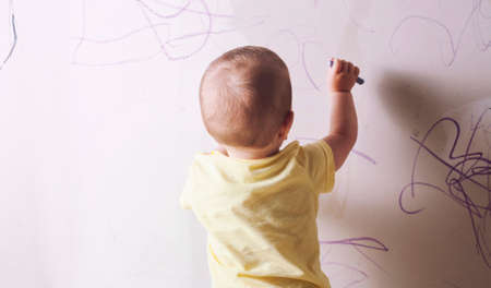 Baby boy drawing with wax crayon on plasterboard wall. He is with his back towardsの写真素材