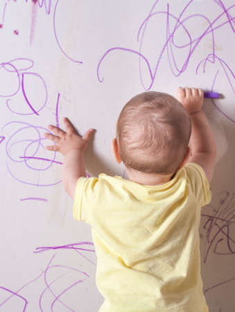 Baby boy drawing with wax crayon on plasterboard wall. He is with his back towards