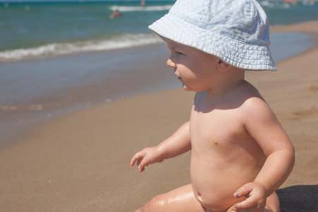 Happy baby boy with blue hat sitting on the sand and observing the seashore on the beachの写真素材