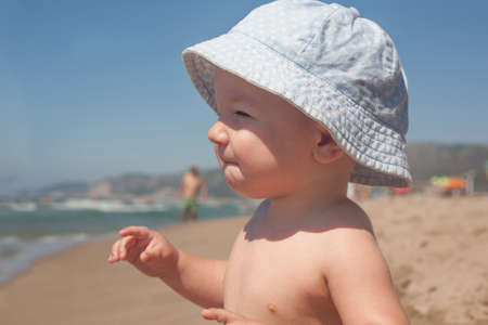 Happy baby boy with blue hat sitting on the sand and observing the seashore on the beachの写真素材