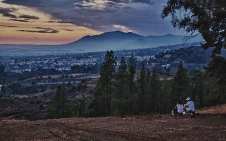 Unrecognizable couple sitting on the top of the hill, observing the sunset with their dog