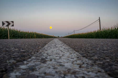 Red car traveling down by corn fields with full moon at dusk. low-angle viewの写真素材