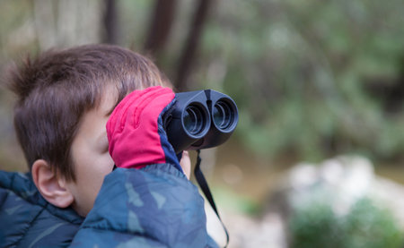 Child boy holds binoculars in nature. He is wearing gloves and a camouflage anorakの写真素材