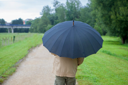 A retired man, protected by an umbrella, walks through a green meadow. Concept of financial protection through retirement pensions.の写真素材