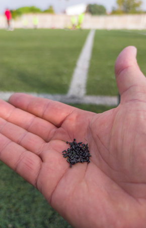 Hand holds rubber pellets from synthetic turf. Soccer field with player as background