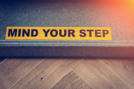 Yellow and black warning sign on stairs stating "Watch your Step" signifying a potential trip hazard, steep stairs and care to be taken when going up or down stairsの写真素材