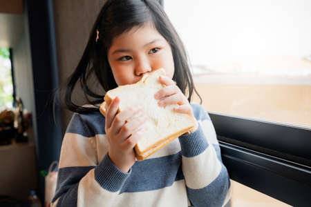 asian healthy kid girl in blue and white sweater enjoy and feel good with bread sandwish in kitchen