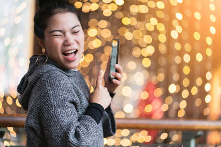 beautiful attractive asian female hand hold smartphone with laugh and smile happiness joyful with background of festive celebrate bokeh lightの写真素材