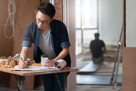 asian attractive glasses interior designer working at house site renovation construction progress with smile and confident at desk with blue print and drawing toolの写真素材