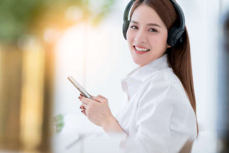 beauty asian female white dress long hair wear headphone and use type search smartphone on white bed with clam and cheerful white bedroom interior backgroundの写真素材