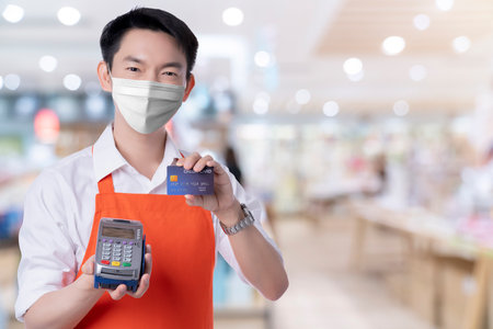 business male owner apron uniform wear surgical protective mask from coronavirus covid-19 spread hand welcome and hold credit card present online purchase payment to avoid virus infected new normalの写真素材