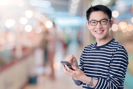 smart casual asian happiness male wear glasses smile and cheerful hand use smartphone ready to press buy button with shopping mall abstract blur background technology communication ideas conceptの写真素材