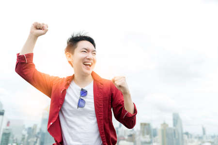 Asian casual businessman wearing red shirt toothly smile wide mouth open excited and cheerful smartphone screen display with capital building cityscape background business successful ideas conceptの写真素材