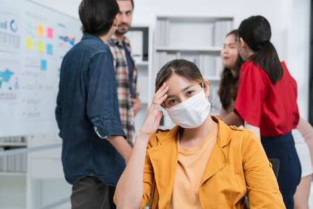 sick asian worker female wear mask with covid19 corona flu disease infection sit and hand touch head in meeting room with background of colleague office friend discussion office baの写真素材