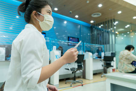 New normal asian female sit and write paper work at commercial bank with face mask and social...
