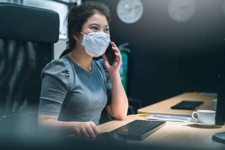 young asian female wear facemask protection while working hard busy multi task with smartphone and table computer late night new normal work lifestyle in officeの写真素材