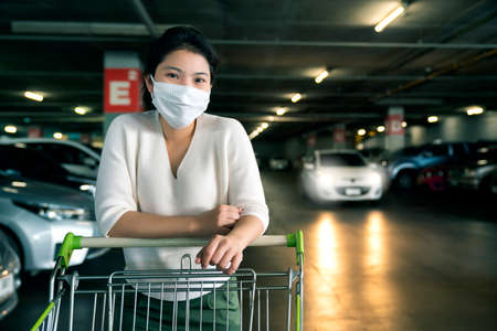 portrait young asian female wear face mask protection hand pull shopping cart alone in parking lot...