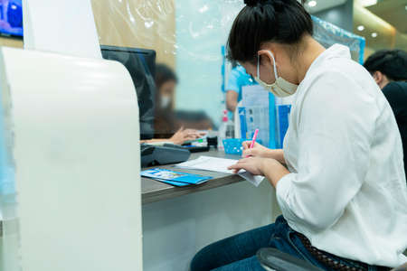 New normal asian female sit and write paper work at commercial bank with face mask and social...