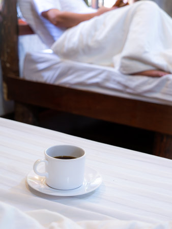 The White Coffee Mug Rests on The White Mattress behind The Man Who Sitting on The Bed Playing a Cell Phoneの写真素材