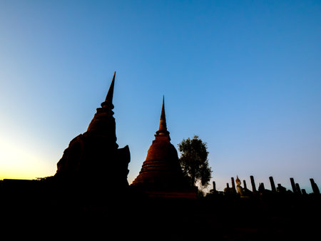 Silhouette of two ancient chedis (stupas) against a vibrant sunset sky at Sukhothai Historical Park, Thailand. In the background, ancient Buddha statues and pillars add to the serene atmosphere of this UNESCO World Heritage site. This image captures the beauty and historical significance of Sukhothai architecture and Buddhist heritageの写真素材