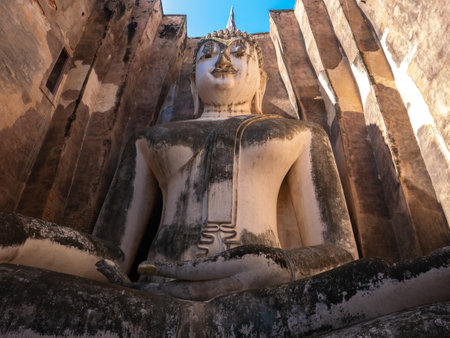 An ant's eye view of a large stucco Buddha image, nearly full figure, known as Phra Achana, within Wat Si Chum in Sukhothai Historical Park. Surrounded by weathered ancient brick walls and a bright blue sky, the image conveys the grandeur, sanctity, and age of this historic siteの写真素材