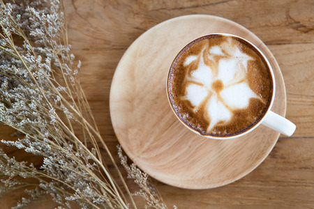 Cup of cappuccino with latte art on a wooden plate and wooded table  decorated with dried glass flower on the sideの写真素材