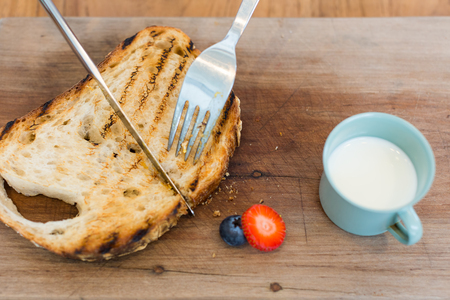 Cutting toasted bread using fork and knife on a wooden plank with slice of fruit and cup of milk on...
