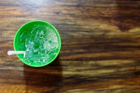 Glass of ice in green plastic cup with a white straw on a wooden table.の写真素材