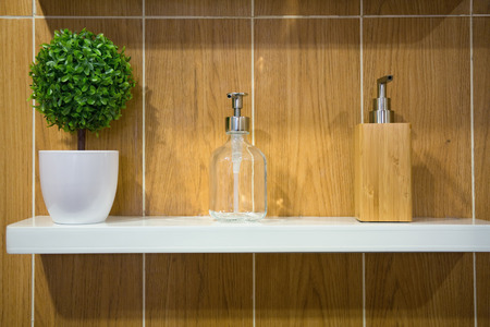 Soap dispensers and small plastic tree in a white ceramic cup on a white shelf.の写真素材