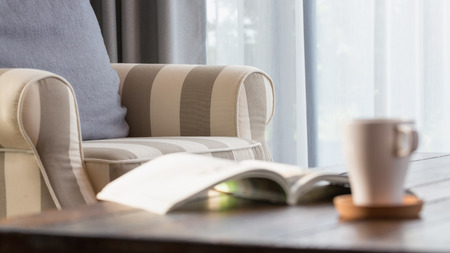 Cozy armchair with gray pillow in a bright room with white curtain , wooden tea table  with an opened book and a cup of coffee.の写真素材
