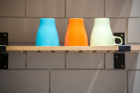 Colorful ceramic coffee cups on a wooden shelf against white kitchen wall.の写真素材