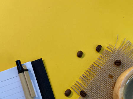 On the yellow background is an open notebook and a pen, next to a jute napkin there is a wooden cup with coffee and sprinkled coffee beansの写真素材