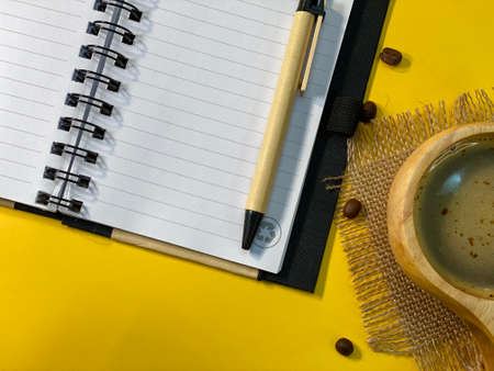 On the yellow background is an open notebook and a pen, next to a jute napkin there is a wooden cup with coffee and sprinkled coffee beansの写真素材