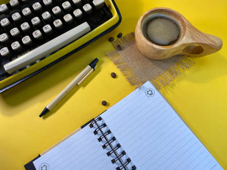 On a yellow background there is a typewriter and a notebook unfolded, next to a jute napkin there is a wooden cup with coffee and sprinkled coffee beansの写真素材