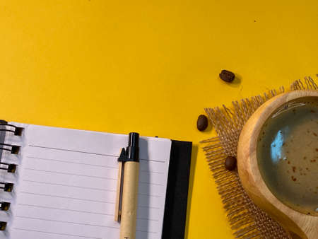 On the yellow background is an open notebook and a pen, next to a jute napkin there is a wooden cup with coffee and sprinkled coffee beansの写真素材