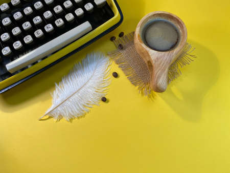 On a yellow background there is a typewriter, next to a jute napkin there is a wooden cup with coffee and sprinkled coffee beansの写真素材