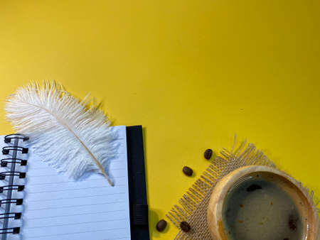 On the yellow background is an open notebook and a pen, next to a jute napkin there is a wooden cup with coffee and sprinkled coffee beansの写真素材