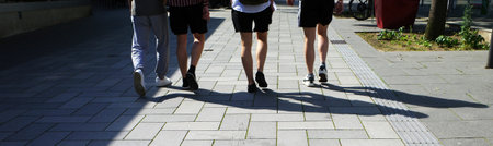 The legs of four male friends. They walk on a broad boulevard. Three of them have bare legs. Their...
