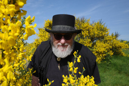 An older man, dressed in black with a modern version of a top hat, looks through blooming common...